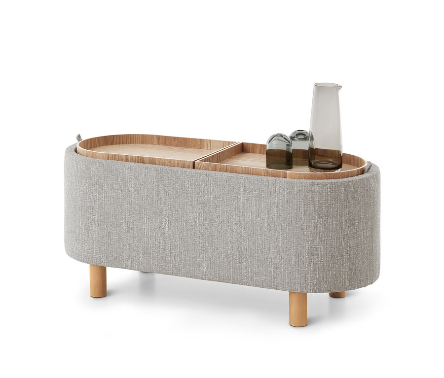Heller, ovaler Stoffhocker mit vier Holzbeinen und zwei abnehmbaren Holztabletts oben. Ein Tablett enthält eine graue Glaskaraffe und zwei passende Gläser.