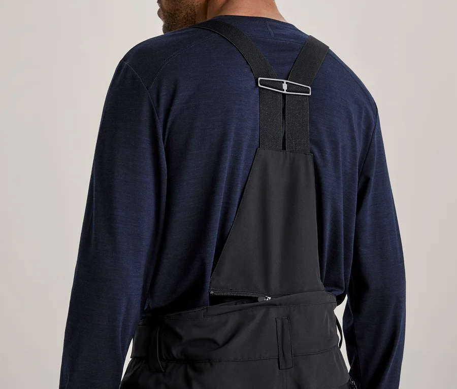 Rückenansicht einer Person in einem dunkelblauen Langarmshirt und schwarzer Latzhose mit Hosenträgern, die sich mit einer silbernen Schnalle treffen.