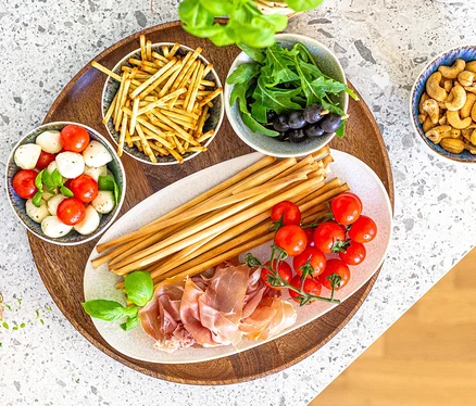 Aufsicht auf eine Vorspeisenplatte auf einem Holztablett, mit einer ovalen Platte aus dem Mäser Platten-Set »Frozen«, 3-tlg, belegt mit Grissini, Prosciutto, Kirschtomaten und Basilikum. Umgeben von Schalen mit Mozzarella-Tomaten, Knabberstangen, Rucola-Oliven und Cashewkernen.