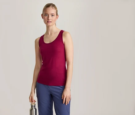 Eine Frau mit zurückgebundenen blonden Haaren, die ein magentafarbenes Tanktop und blaue Hosen trägt, steht nach vorne gerichtet und hält eine silberne Wasserflasche in ihrer rechten Hand.