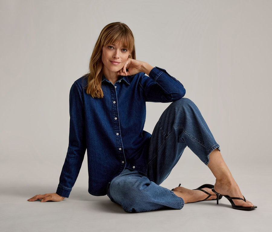 Frau sitzt auf dem Boden, trägt ein dunkelblaues Jeanshemd, hellblaue Cropped-Jeans und schwarze Sandalen mit hohen Absätzen, blickt in die Kamera und stützt ihr Kinn ab.