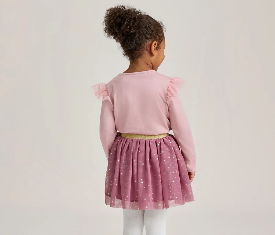 Junges Kind von hinten, trägt ein rosa Langarmshirt mit Rüschen an den Schultern, einen lila Tüllrock mit goldenen Sternen und einen goldenen Bund sowie weiße Strumpfhosen.