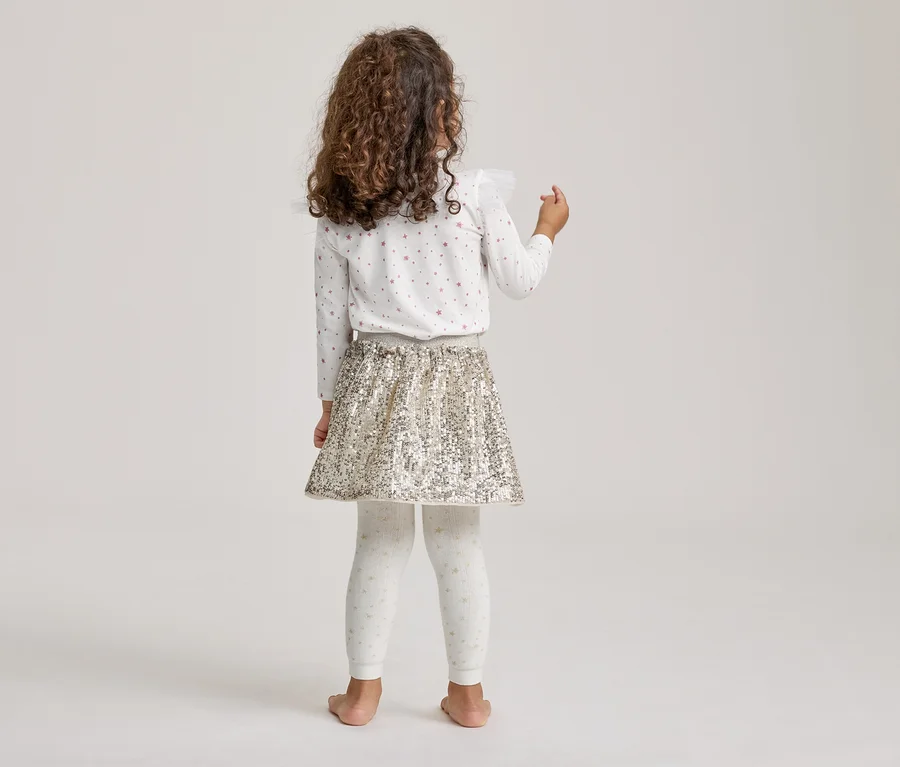 Kind steht mit dem Rücken zur Kamera, trägt ein weißes Langarmshirt mit Sternen, einen goldenen Paillettenrock und weiße gemusterte Leggings.