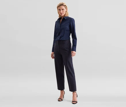 Blonde Frau steht in einem dunkelblauen Langarmhemd, einer dunkelblauen Cropped-Hose und schwarzen Sandaletten mit hohen Absätzen.