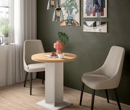 Runder Holztisch mit weißem Sockel und zwei beigen Stühlen in einem Raum mit dunkelgrüner Wand. Auf dem Tisch stehen eine korallenfarbene Vase, eine Tasse und Macarons. Über dem Tisch hängen zwei Glaspendelleuchten. An der Wand hängen drei gerahmte Bilder.