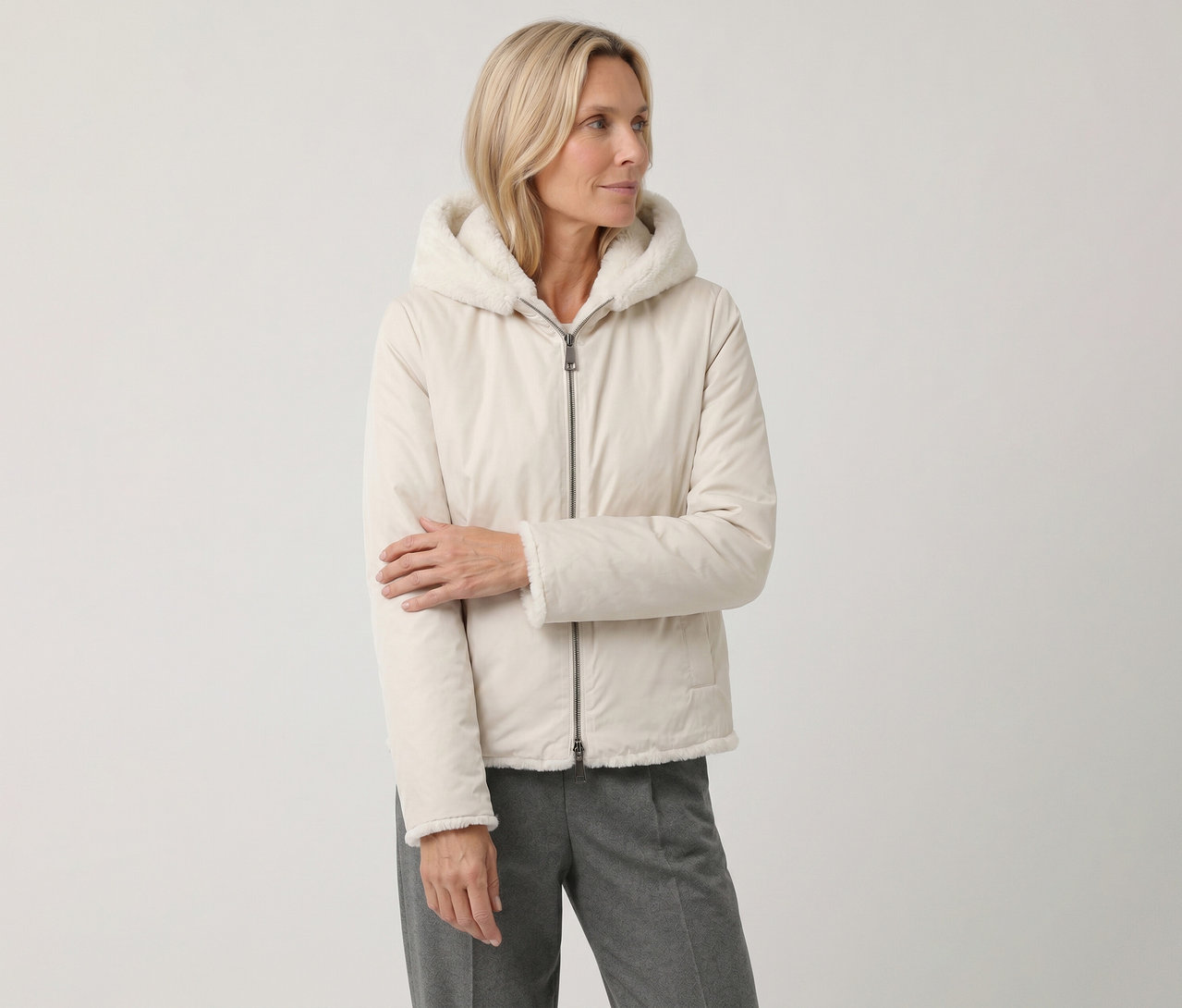 Blonde Frau trägt eine hellbeige Kapuzenjacke mit flauschigem weißem Futter und graue Hose, blickt nach rechts.