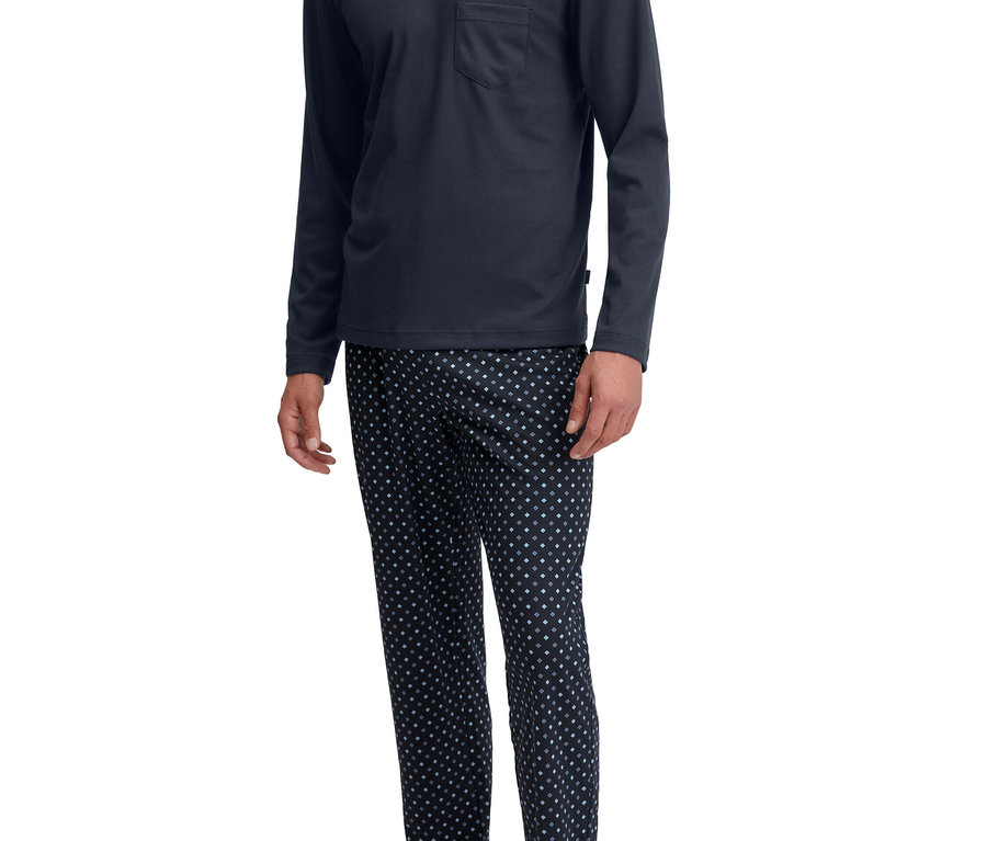 Mann steht lächelnd in einem dunkelblauen Langarm-V-Ausschnitt-Pyjamaoberteil mit Brusttasche und dunkel gemusterter Pyjamahose, barfuß.