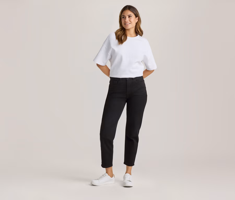 Stehende Frau in weißem Oversize-T-Shirt, schwarzer Cropped-Jeans und weißen Sneakern, mit Händen hinter dem Rücken lächelnd.