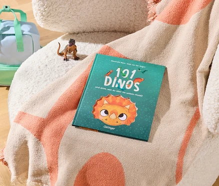 Ein hellblauer und mintgrüner Rucksack steht auf einem Holzboden neben einem cremefarbenen Sessel. Auf dem Sessel liegt eine beige-orange Decke mit einem kleinen braunen Dinosaurier-Spielzeug und einem türkisfarbenen Buch mit dem Titel „101 DINOS“.