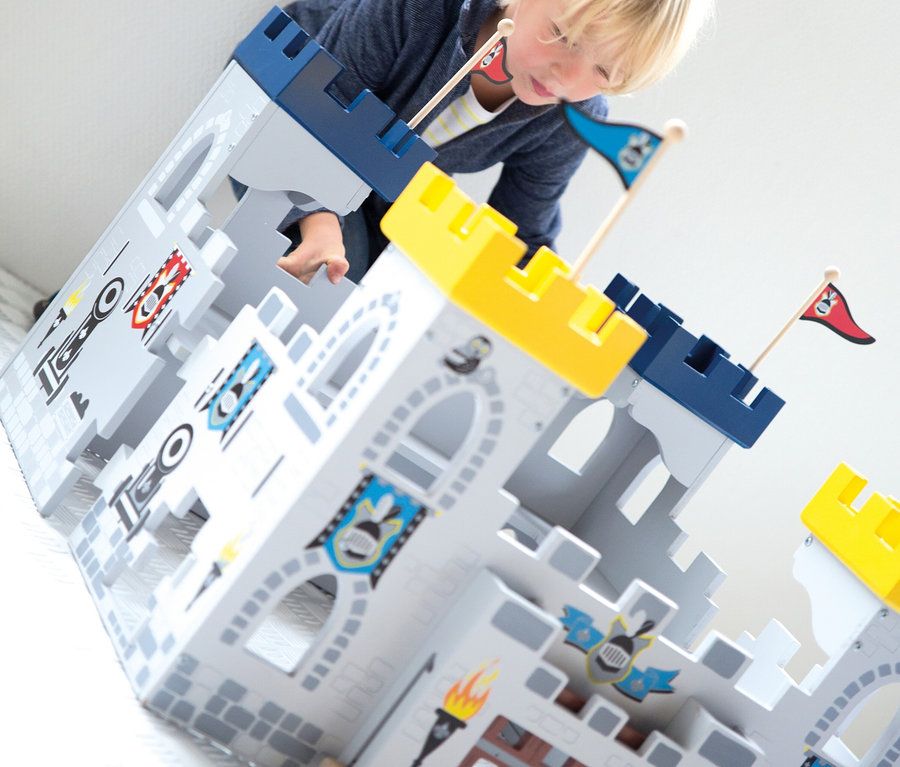 Junger blonder Junge beugt sich über ein graues Holzspielzeugschloss mit blauen und gelben Zinnen, Fahnen und Schilden und spielt mit den Händen darin.