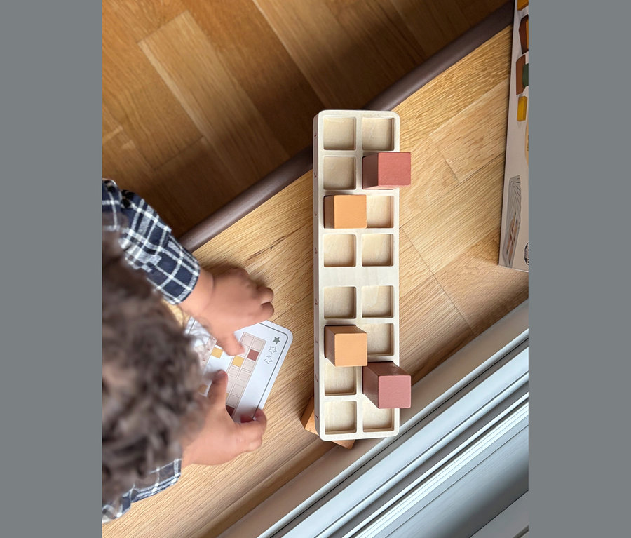 Kinderhände halten eine Musterkarte neben einem Holzspielzeug mit orangefarbenen und rotbraunen Blöcken auf einem Holzboden.