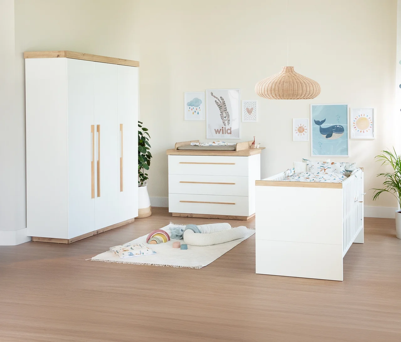 Helles Kinderzimmer mit weißem Kleiderschrank, Wickelkommode und Gitterbett mit Holzdetails. An der Wand hängen bunte Bilder, auf dem Boden liegt ein Teppich mit Babyspielzeug.
