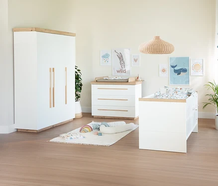 Helles Kinderzimmer mit weißem Kleiderschrank, Wickelkommode und Gitterbett mit Holzdetails. An der Wand hängen bunte Bilder, auf dem Boden liegt ein Teppich mit Babyspielzeug.