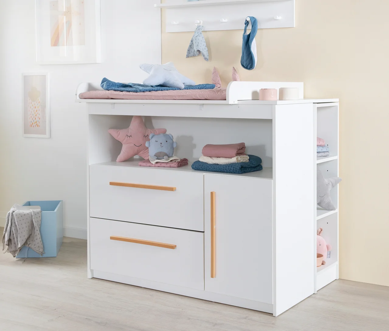 Weißer Wickeltisch mit Wickelauflage, blauer und rosa Decke, Sternenkissen und Einhornkissen. Darunter Schubladen und offene Fächer mit Babykleidung und Spielzeug. Links ein blauer Aufbewahrungskorb, an der Wand Bilder und ein Regal mit Haken.