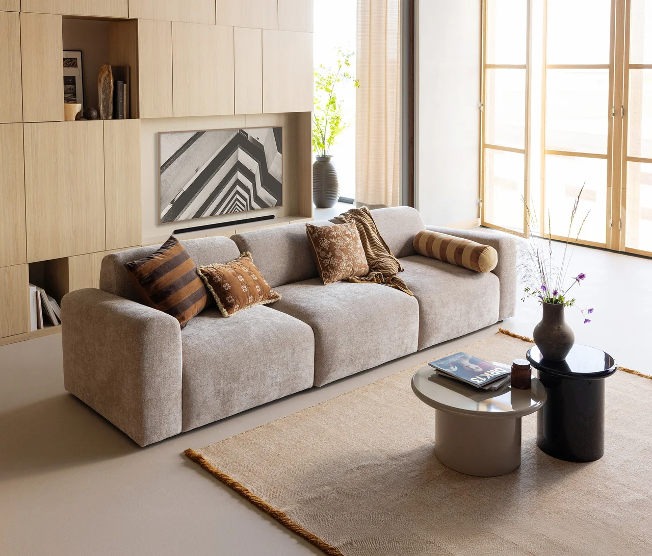 Modernes Wohnzimmer mit hellem Modulsofa, braunen Kissen und Decke. Davor liegt der hellbraune WOOOD Teppich »Weavly«, ca. 170 x 240 cm, und zwei Couchtische. An der Wand ist ein TV in einer Holzschrankwand.
