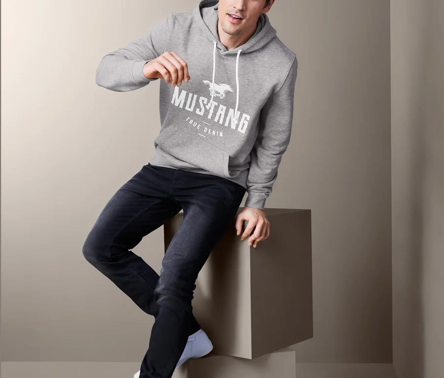 Mann sitzt auf einem Würfel und trägt einen grauen Mustang Hoodie, Jeans und Turnschuhe.