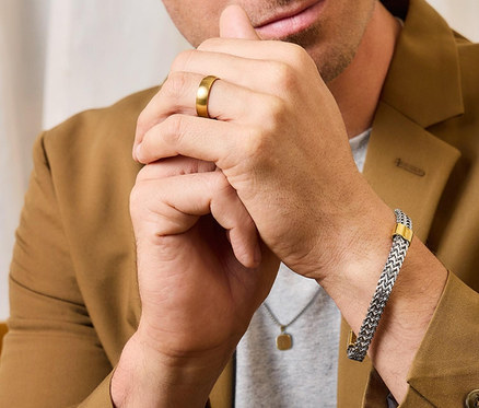 Mann mit goldenem Ring am Finger und silbernem Armband am Handgelenk, trägt ein braunes Jackett.