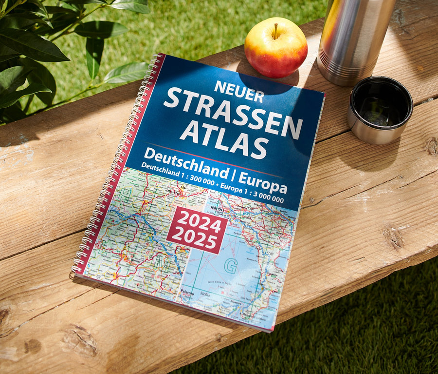 Auf einem Holztisch liegen ein Apfel, eine Thermoskanne und das Buch: Neuer Straßen-Atlas Deutschland/Europa 2024/2025.