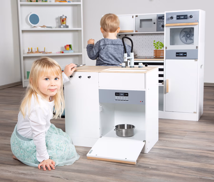 Blondes Mädchen kniet neben einer weißen Spielküche. Ein Junge steht an der Spüle der small foot Kinderküche modular XL.