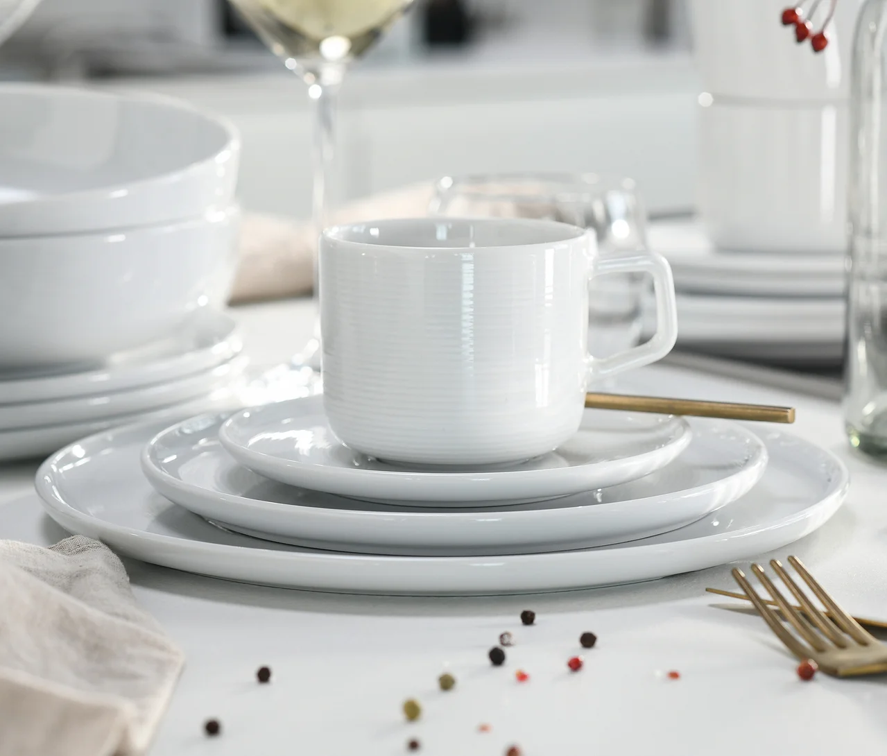 Weißes creaTable Service "Session White" Geschirrset auf einem Tisch, mit einer gerippten Tasse auf gestapelten Tellern, Schüsseln, einer goldenen Gabel und verstreuten Pfefferkörnern.