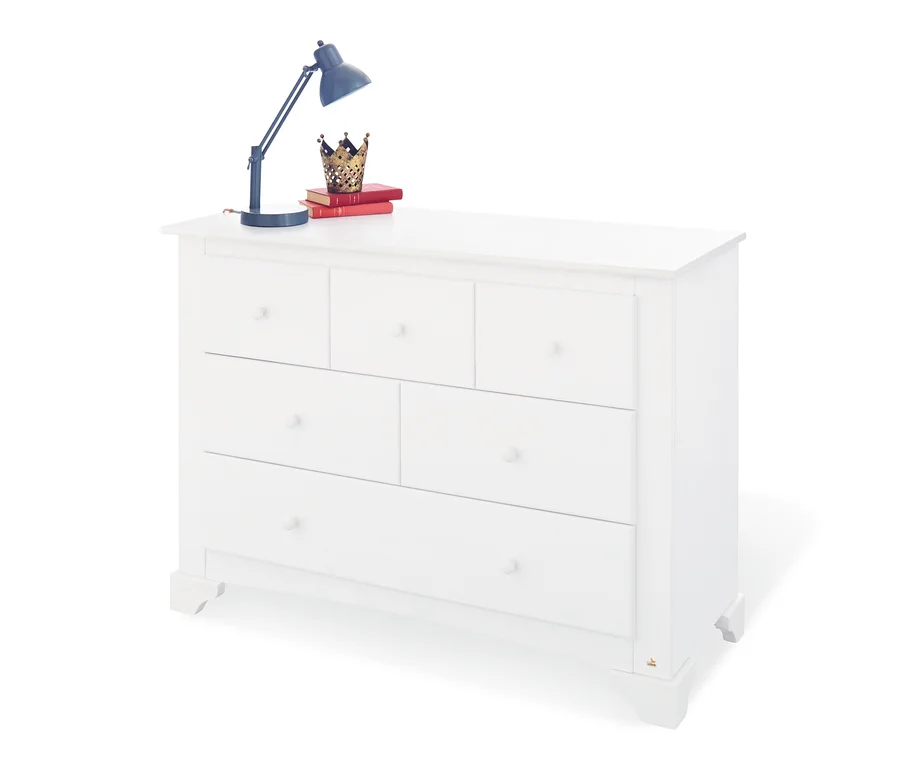 Weiße Kommode mit sechs Schubladen, auf der ein Buch, eine Krone und eine Lampe liegen.