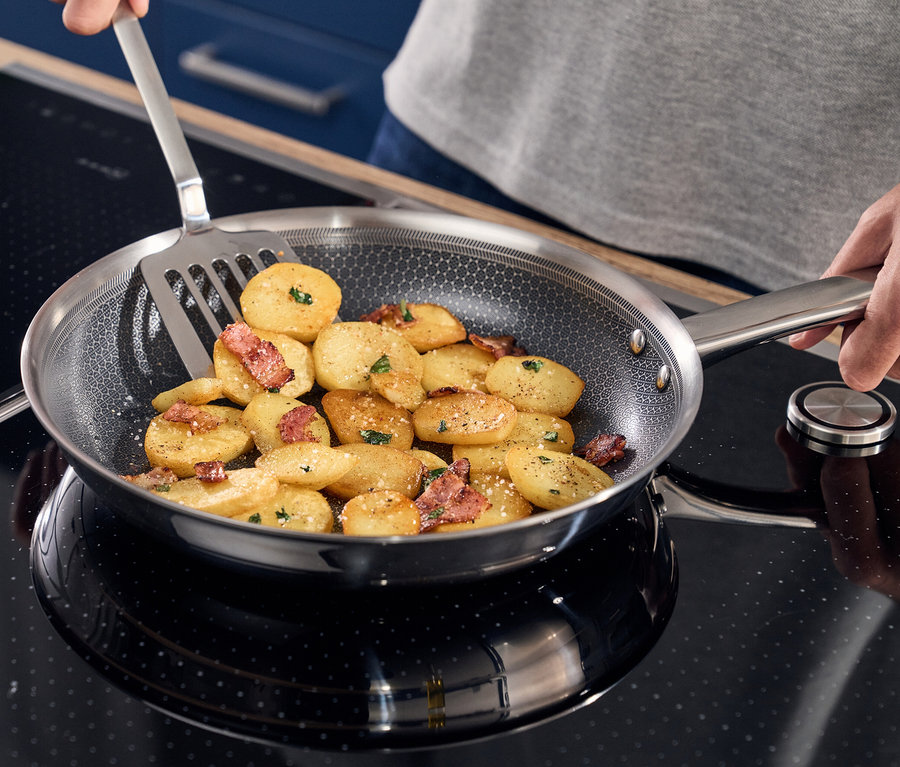 Eine Person brät Kartoffeln mit Speck in einer Henckels by ZWILLING »Valley« Bratpfanne auf einem Induktionskochfeld und verwendet dabei einen Pfannenwender.