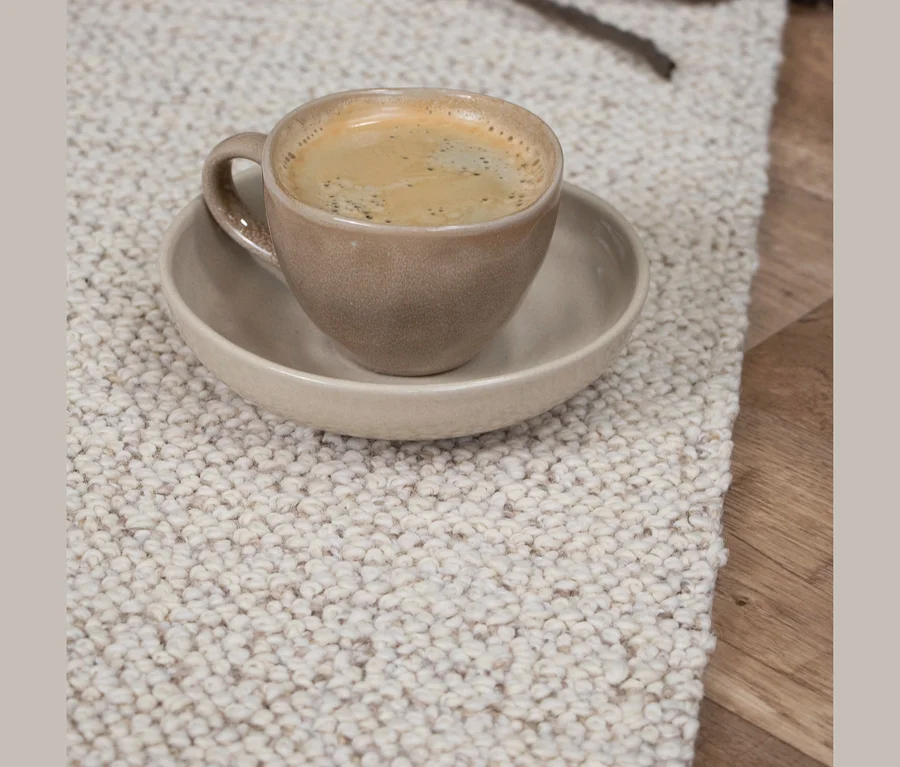 Eine beige Tasse mit Kaffee und Schaum steht auf einer passenden Untertasse auf einem hellen, strukturierten Teppich neben einem Holzboden.