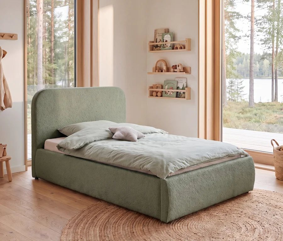 Kinderzimmer mit grünem, texturiertem Einzelbett, hellgrüner Bettwäsche und Sternkissen. Holzregale zeigen Bücher und Holzspielzeug. Große Fenster blicken auf Wald und See; ein runder Flechtteppich liegt auf dem Holzboden.