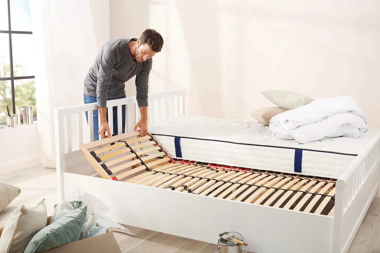 Mann legt Lattenrost in ein weißes Bett ein. Auf dem Bett liegen eine Matratze und eine gefaltete Decke.