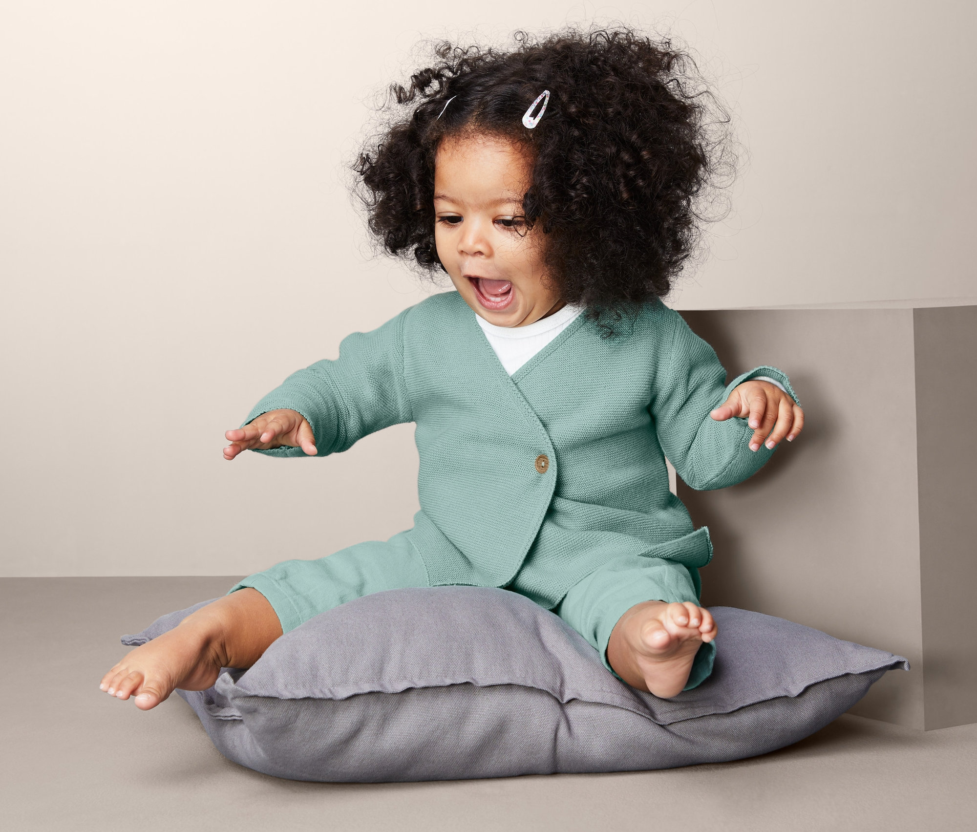 Kleinkind sitzt auf einem Kissen und trägt eine Wickeljacke.