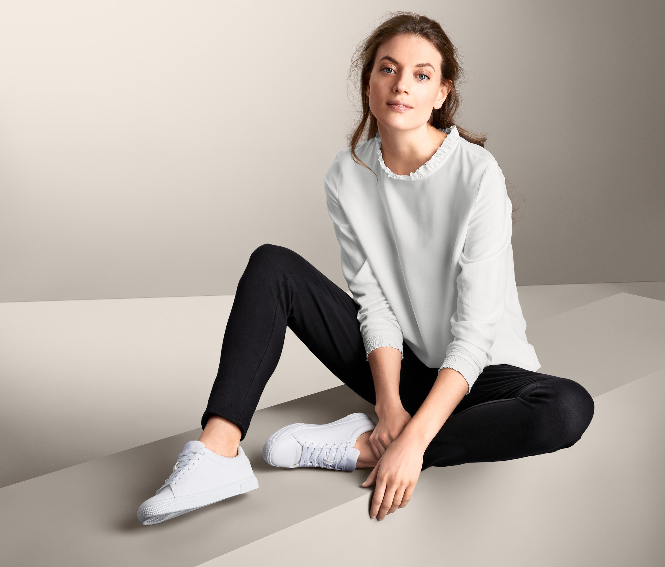 Eine Frau sitzt und trägt eine Bluse, schwarze Hosen und weiße Turnschuhe.