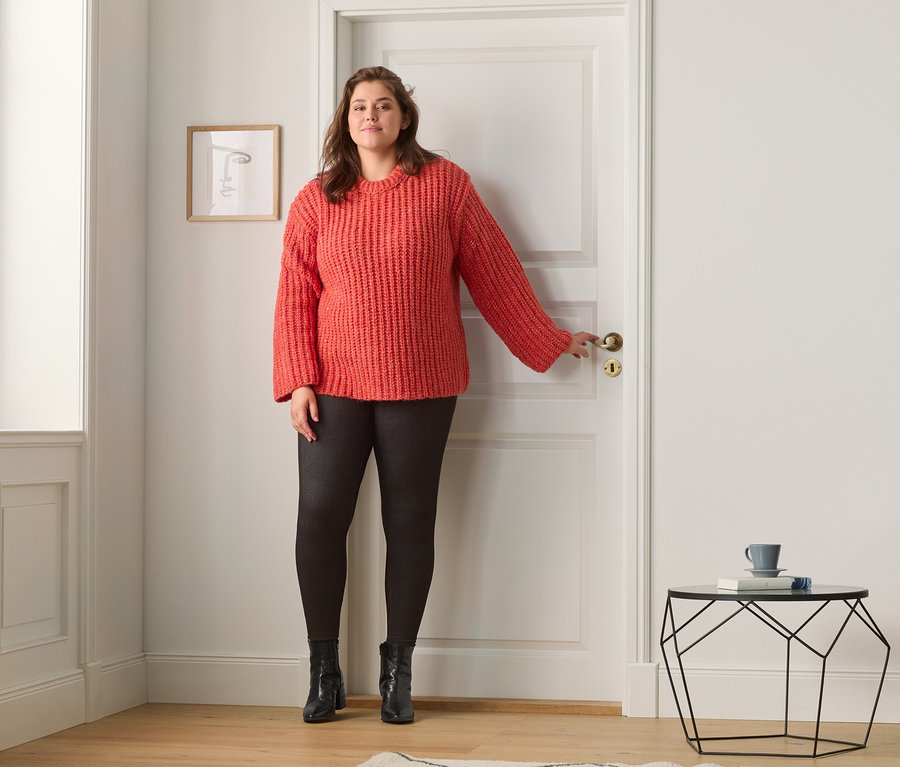 Eine Frau steht an einer Tür und trägt eine gefütterte Leggings im Jeans-Look und einen Grobstrickpullover.