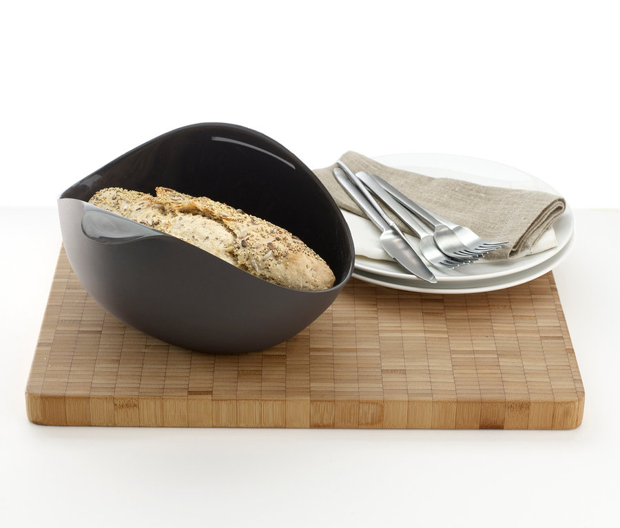 Brot im schwarzen Lékué Brotback-Set auf Holzbrett, daneben Teller mit Serviette und Besteck.