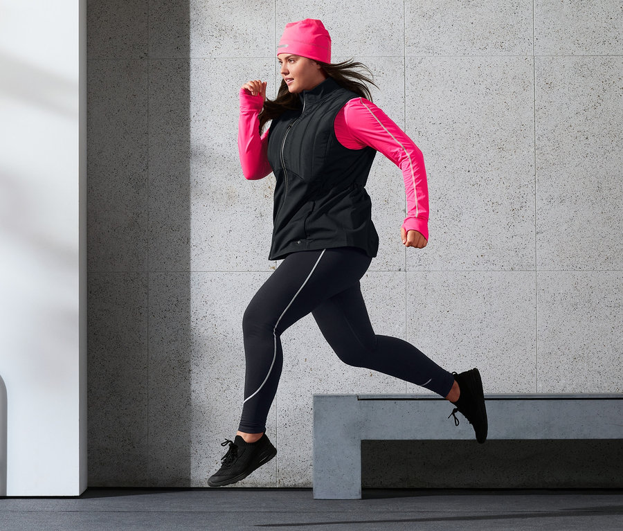 Frau läuft in Thermo-Steppweste, Sporttight, Thermomütze mit Reflektorprint und neonpinkem Langarm-Funktionsshirt.