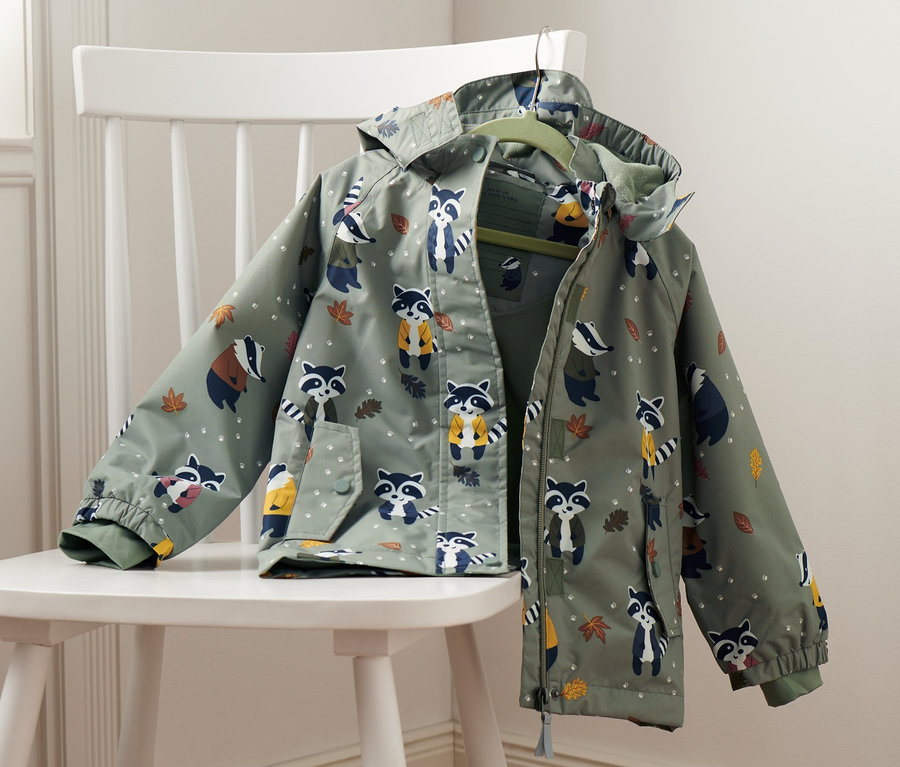 Eine grüne Thermo-Regenjacke mit Dachsprint hängt auf einem weißen Stuhl neben grünen Gummistiefeln.