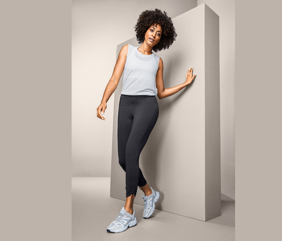 Frau posiert in 7/8-Sporttights, weißem Tanktop und Turnschuhen.