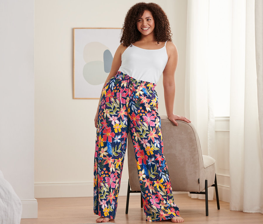 Eine Frau, die sich an einen Stuhl lehnt, trägt ein weißes Hemdchen und eine dunkelblaue Relaxhose mit Blumenmuster.