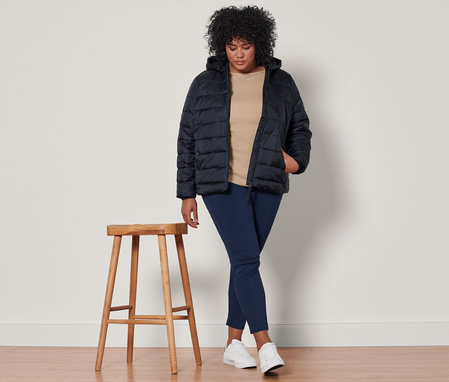 Eine Frau steht und trägt eine dunkelblaue Steppjacke mit Kapuze, ein beiges Shirt mit 3/4-Arm und eine dunkelblaue Stretchhose.