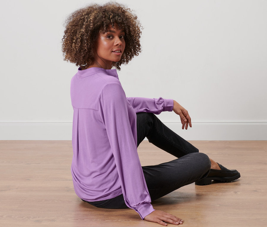 Frau sitzt auf dem Boden und trägt ein fliederfarbenes Blusenshirt mit Knopfleiste.