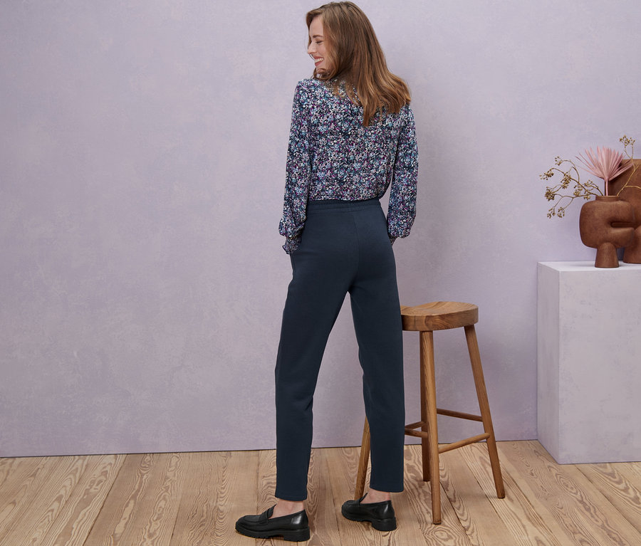 Frau steht mit dem Rücken zur Kamera und trägt eine Sweathose, eine geblümte Bluse und schwarze Schuhe.