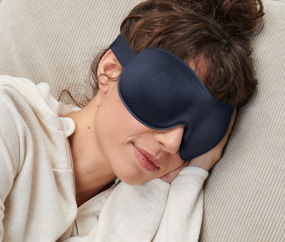 Frau schläft mit einer Schlafmaske auf einer Couch.