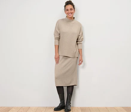 Eine Frau steht und trägt einen Feinstrickpullover mit Stehkragen und einen Strickrock.