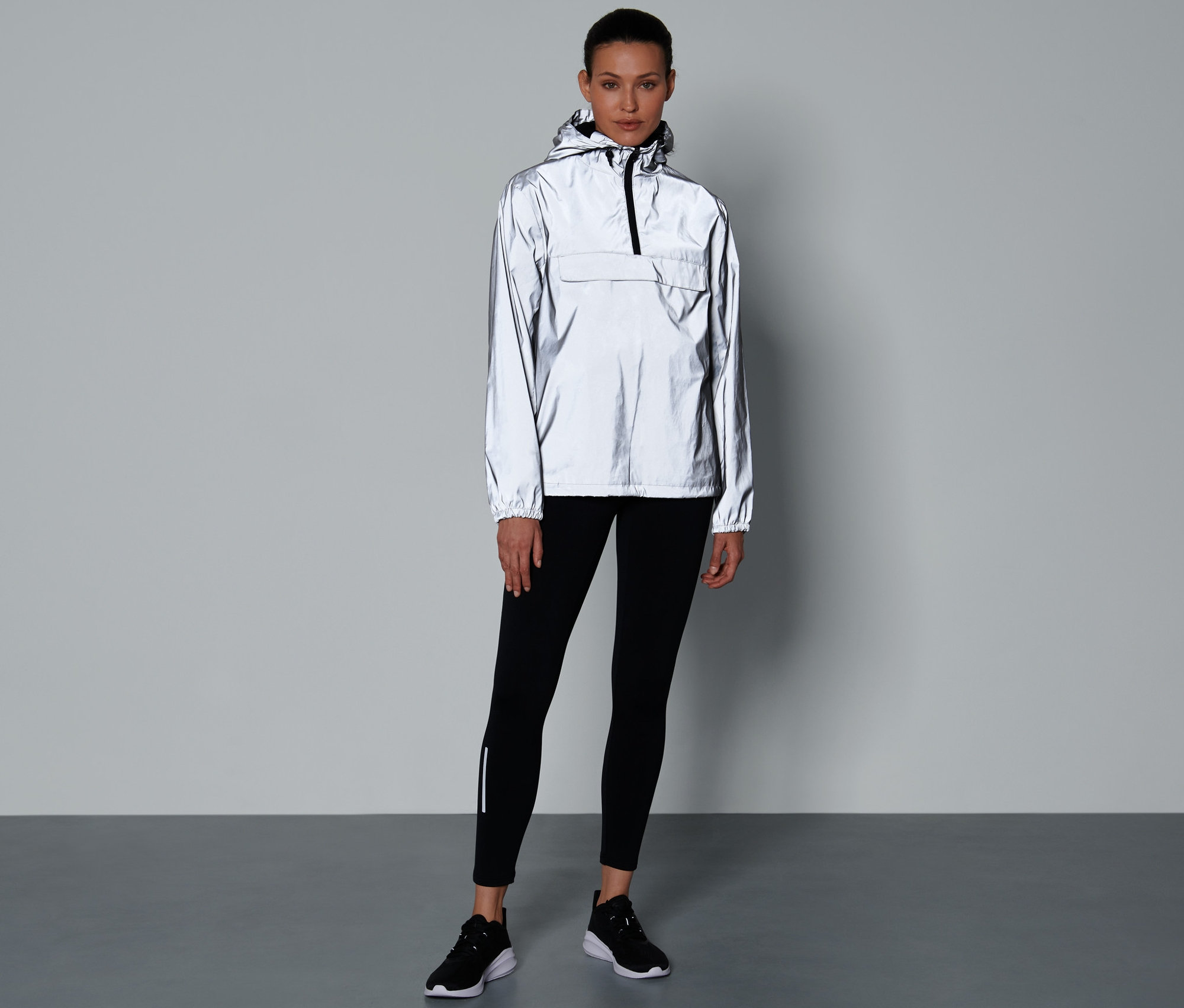 Frau steht und trägt einen reflektierenden Windbreaker, schwarze Leggings und Schuhe.