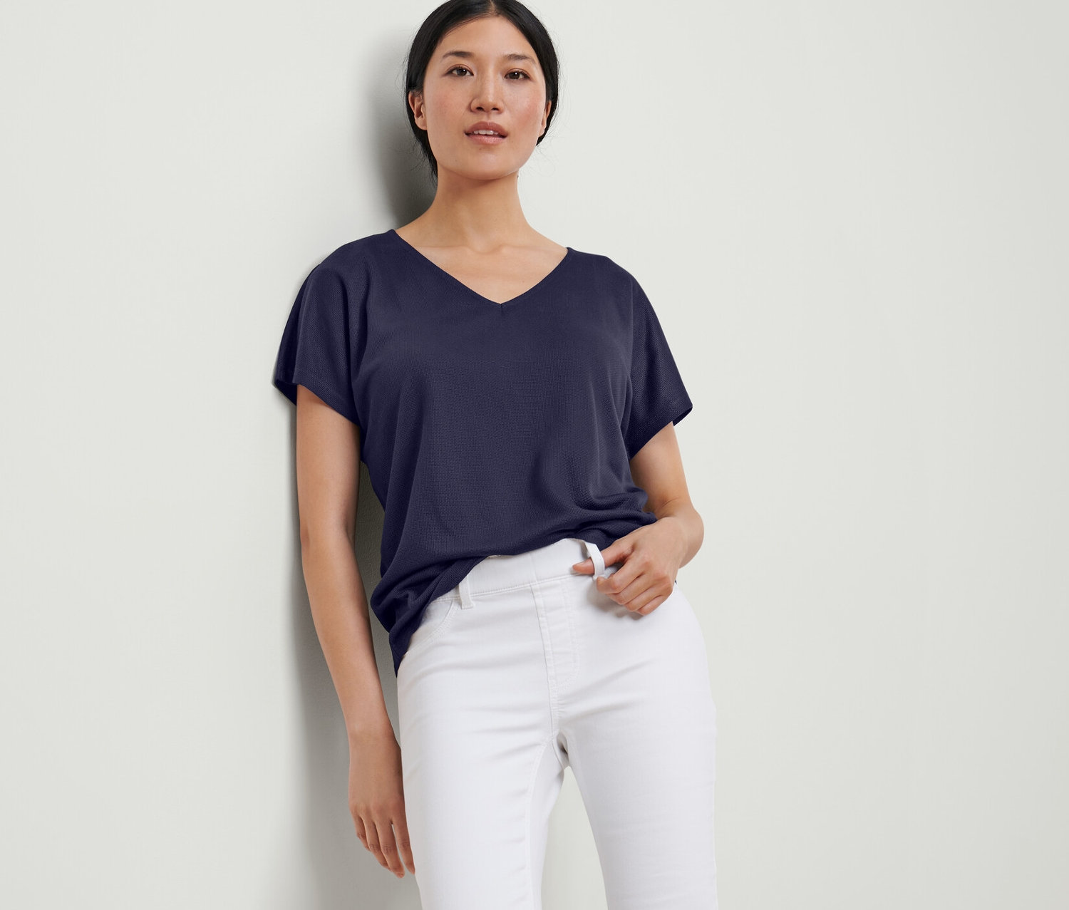 Eine Frau lehnt an einer Wand und trägt ein dunkelblaues, strukturiertes Shirt und eine weiße Hose.