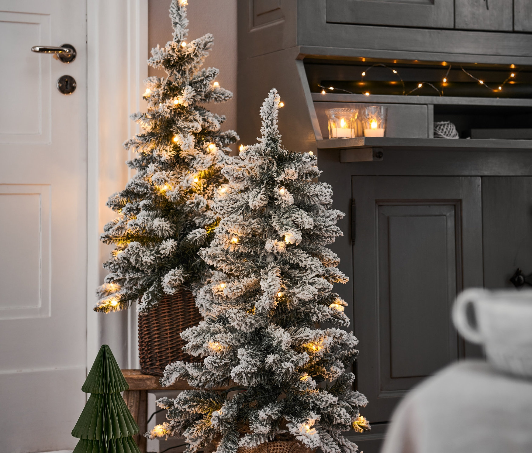 Zwei kleine, beschneite Weihnachtsbäume mit LED-Lichtern stehen auf einem Holzboden. Ein Baum befindet sich in einem geflochtenen Korb, der andere in einem Jutetopf.