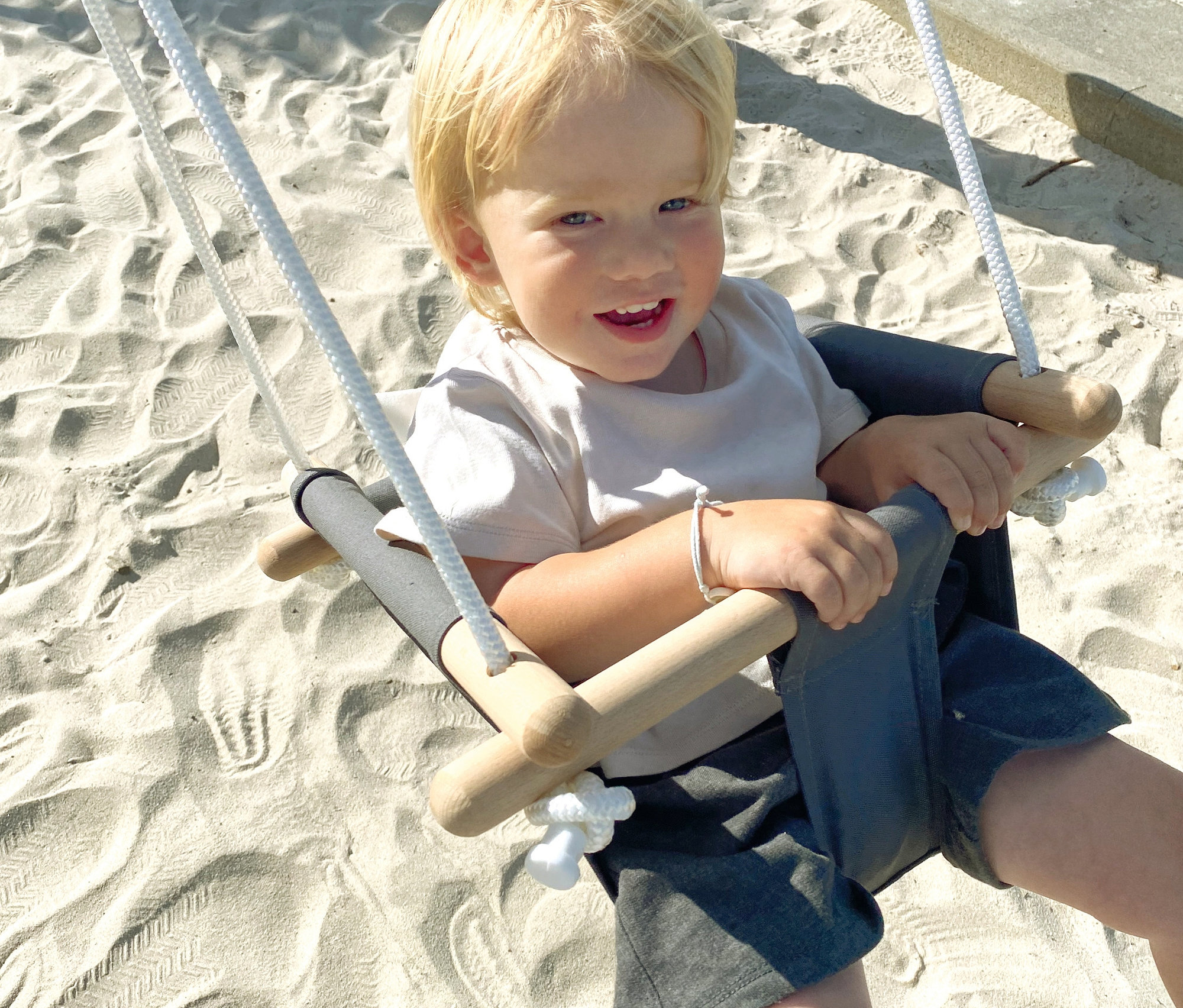 Ein blonder Junge sitzt auf einer small foot Babyschaukel »Komfort« auf einem Sandkasten.