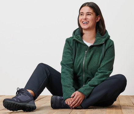 Frau sitzt auf Holzboden und trägt eine grün melierte Kapuzen-Strickfleecejacke und eine Thermohose.