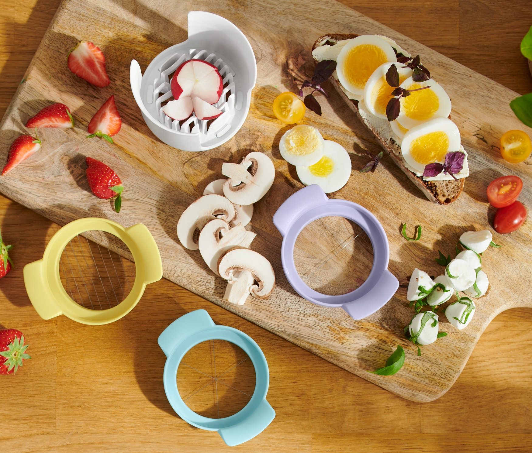 Auf einem Holzbrett liegen Erdbeeren, Pilze, ein 3-in-1-Eierschneider, ein Toast mit Ei, Mozzarella und Cherrytomaten.