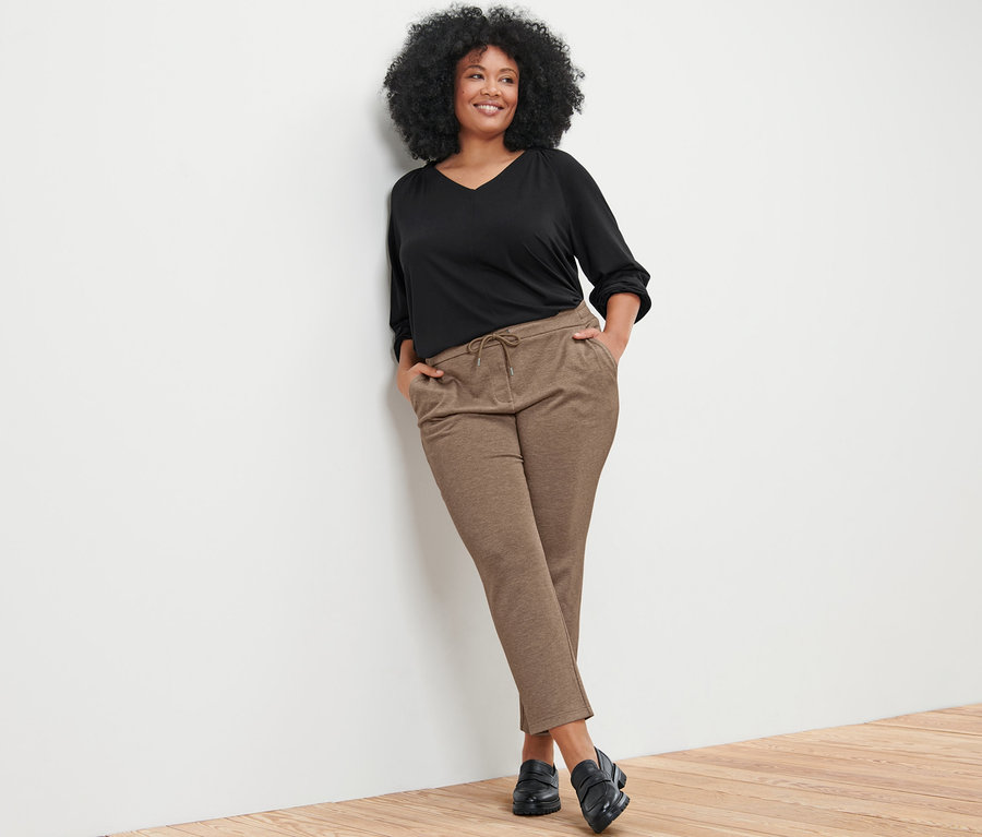 Frau lehnt an einer Wand und trägt ein Blusenshirt mit Raffung und eine beige Joggpants.