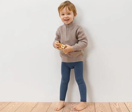 Ein Junge steht an einer Wand und spielt mit einem Holzspielzeug. Er trägt blaue Strickleggings und einen braunen Pullover. 3 Kinder-Strickleggings.